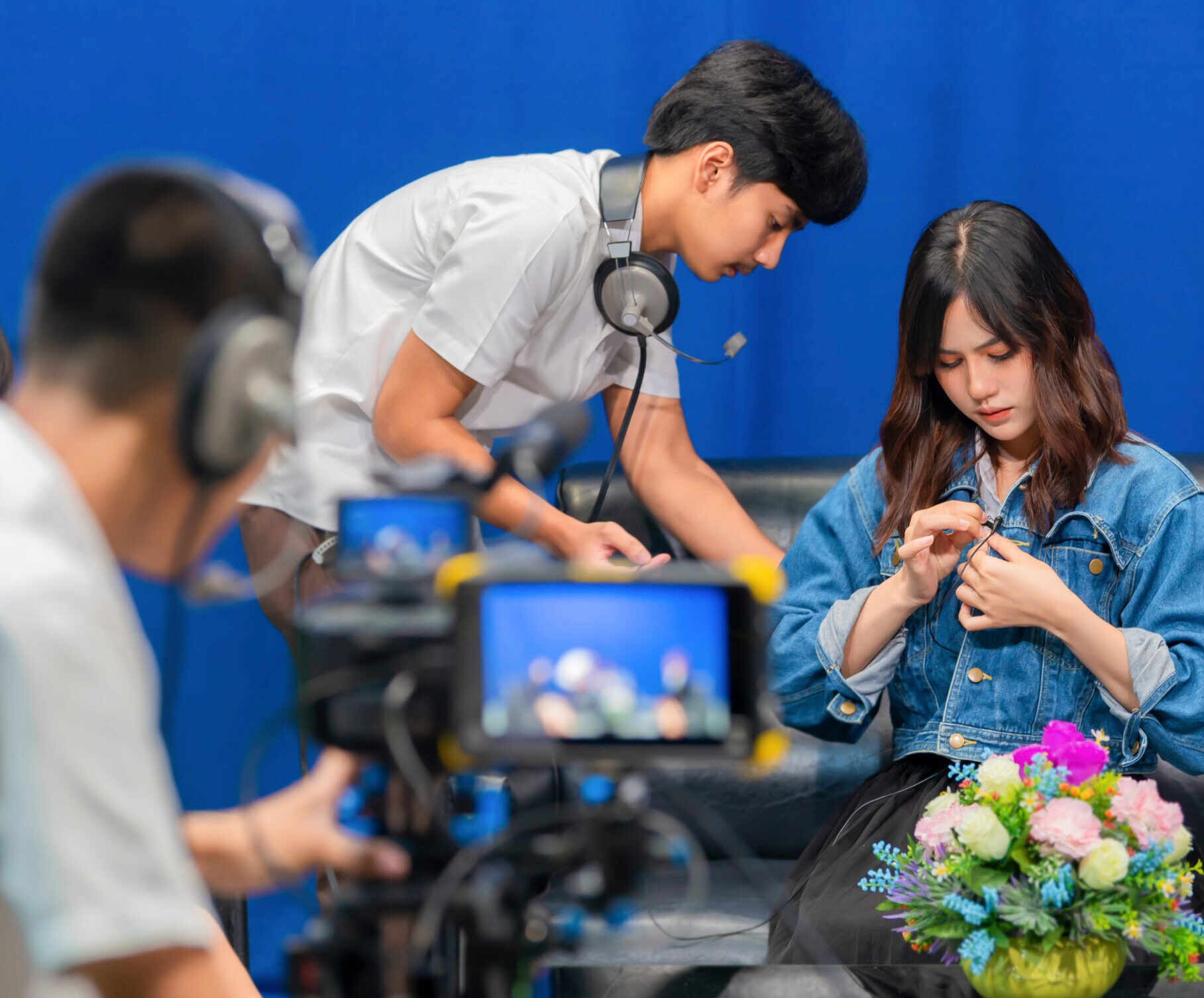 Television video camera recording interview in broadcast studio. Asian woman clipping tie-clip microphone and Professional cameraman with headphones looking video camera viewfinder. Selective focus.