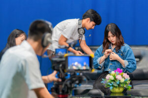 Television video camera recording interview in broadcast studio. Asian woman clipping tie-clip microphone and Professional cameraman with headphones looking video camera viewfinder. Selective focus